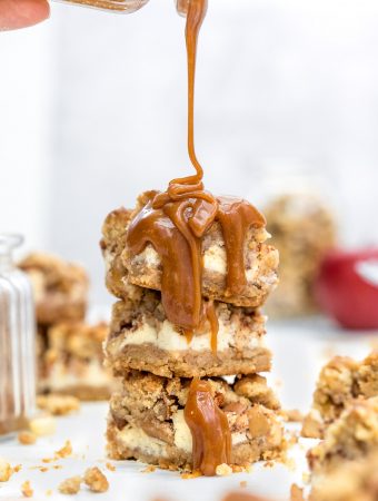 Apple Crumble Cheesecake Bars with Salted Caramel Sauce in a tower stack being poured over with caramel sauce
