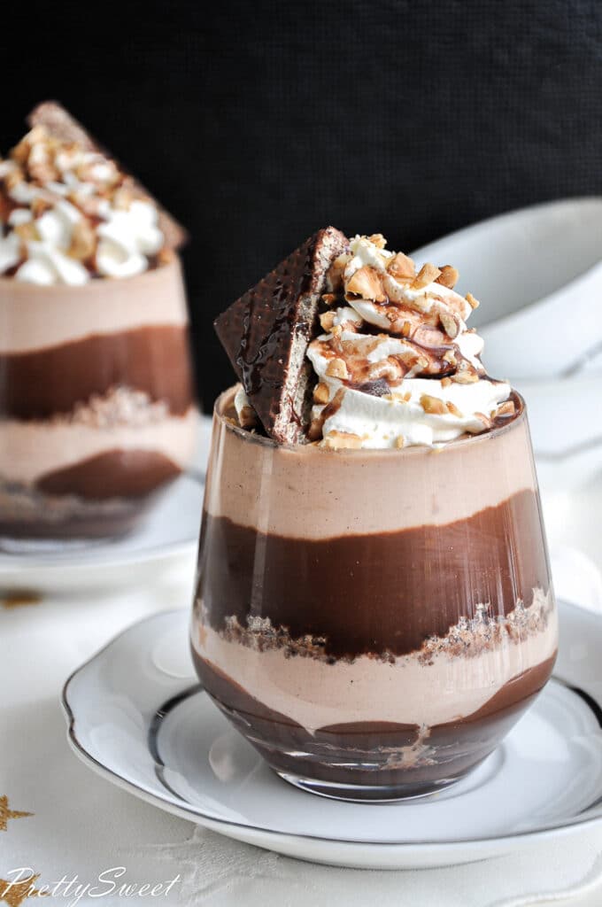 a dessert in a glass with layers of chocolate and mousse with whipped cream on top and another glass in the back