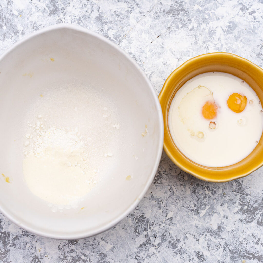 flour with baking powder in a white bowl and eggs with milk in a smaller yellow bowl
